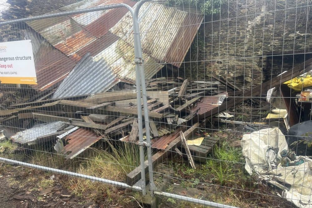 Before: collapsed corrugated-iron outbuilding behind temporary fencing, leaning against the stone wall